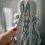 Hand hält ein Schnitzwerkzeug an einer kunstvoll geschnitzten weißen Kerze mit mehrfarbigen Verzierungen