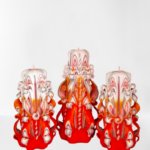 Drei handgeschnitzte Devela Kerzen im 3er-Set, sechseckig und kegelförmig, in warmen Orange- und Rottönen mit filigranen Ornamenten. Eine Kerze ist 21 cm hoch, zwei sind 18 cm hoch – ideal als Herbst-Deko oder luxuriöses Geschenk