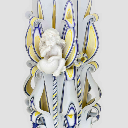 Große handgefertigte weiße Kerze mit zentraler weißer Engelfigur, umgeben von gelb-blauen floralen Schnitzmustern vor hellem Hintergrund