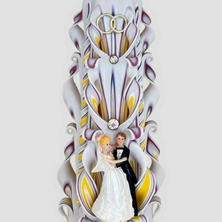Handgeschnitzte weiße Hochzeitskerze mit goldenen und lila Akzenten, goldenen Eheringen, Strasssteinen und Brautpaar-Figur (Braut in weißem Kleid, Bräutigam in Anzug) vor hellem Hintergrund.