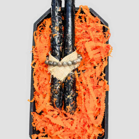 Zwei gekreuzte schwarze handgetauchte Kerzen mit Blütenblättern und Juteschnur vor hellem Hintergrund. Auf den Kerzen Liegt ein Armband aus Edelsteinen
