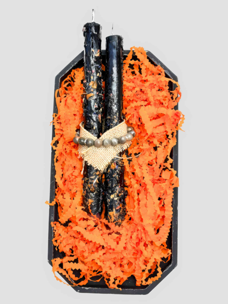 Zwei gekreuzte schwarze handgetauchte Kerzen mit Blütenblättern und Juteschnur vor hellem Hintergrund. Auf den Kerzen Liegt ein Armband aus Edelsteinen