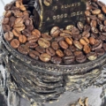 Handgefertigte Kaffeehaus Art-Kerze „Barista“ aus schwarzem Paraffin mit echten Kaffeebohnen, goldener Kaffeetasse und handmodellierten Vintage-Ornamenten. Schild mit Beschriftung:" Coffee is always a good idea"
